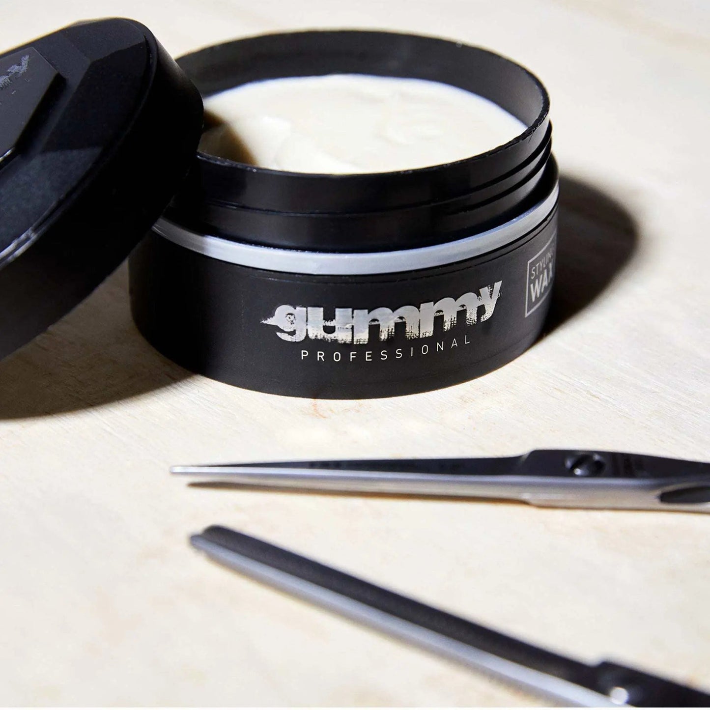 GUMMY STYLING WAX 150 ML, open container showing creamy texture, scissors nearby on wooden surface.